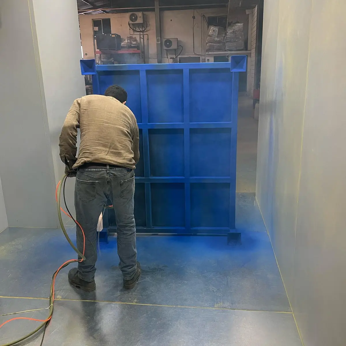 Operador aplicando pintura electrostática en polvo azul sobre una estructura metálica dentro de cabina en Steel Paint